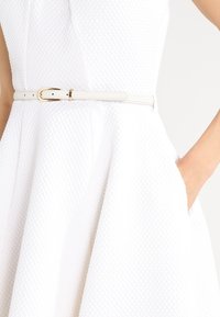 Robe blanche en tissu texturé et matelassé, avec un corsage ajusté, une jupe évasée et une fine ceinture blanche avec une boucle dorée.