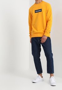 Felpa gialla con logo "Unreleased" in nero, abbinata a jeans cropped blu scuro e sneakers bianche. Tessuto morbido, vestibilità casual.