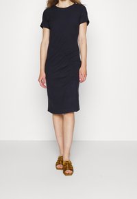 Svart knä-lång klänning med korta ärmar och rynkad sidodetalj, tillverkad i ett mjukt tyg. Matchad med bruna sandaler med remmar.