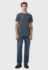 Camiseta de manga corta en gris oscuro con cuello redondo, combinada con jeans de denim azul. El modelo lleva zapatos oscuros y tiene un tatuaje en un brazo.