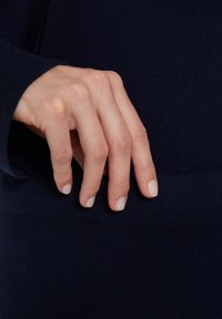 Dunkelblaues, gestricktes Kleidungsstück mit einer Nahaufnahme einer Hand, die auf dem strukturierten Stoff ruht. Glatte, helle Haut mit sorgfältig manikürten Nägeln.