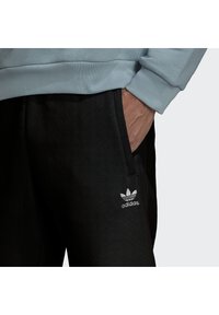 Černé tepláky vyrobené z měkké tkaniny, s bočními kapsami a bílým logem Adidas na levém stehně. Pas je elastický.