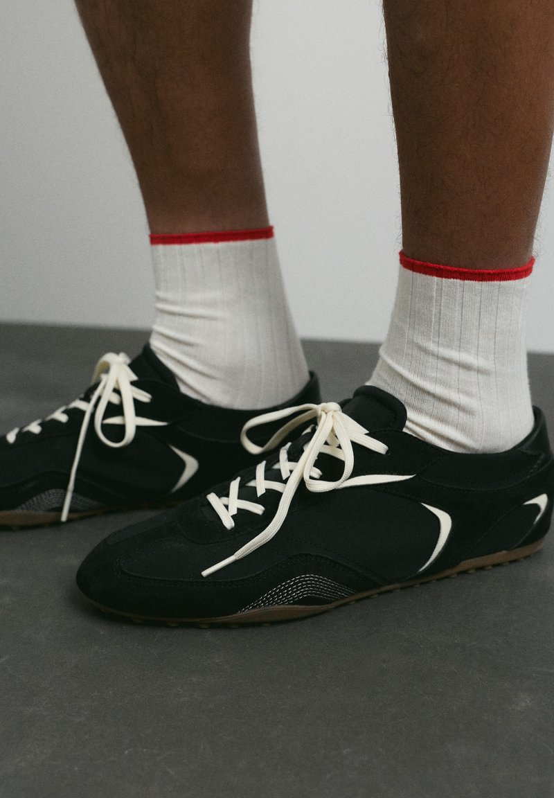 Pieds portant des chaussettes blanches bordées de rouge et des chaussures de sport noires avec des lacets blancs, debout sur un sol sombre.