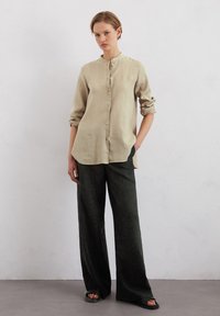 Light beige linen shirt with a mandarin collar, button front, and long sleeves, paired with dark green wide-leg trousers. Sandals visible.