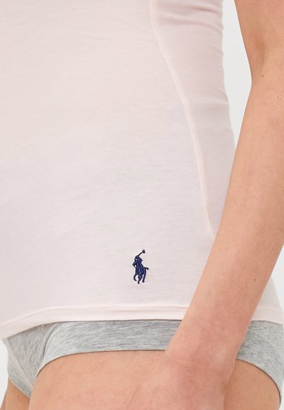 Gaiša rozā pieguļoša T-krekla augšdaļa ar gludu tekstūru, kurai tuvumā ir mazs, tumši zils izšūts logos pie apakšmalas. Saskaņota ar pelēkiem apakšveļas biksēm.