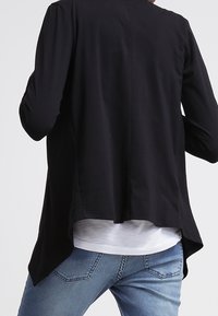 Svart långärmad topp med hög-låg nederkant, med en mjuk textur, minimalistisk design och rundad halsringning. Matchad med ljusblå jeans.