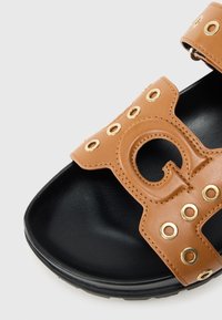 Sandale en cuir marron avec un motif perforé, des œillets dorés et une semelle en caoutchouc noire. Comprend une découpe ornée sur la bride.