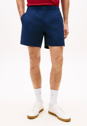 Männliches Model trägt marineblaue, strukturierte Shorts, weiße Socken, weiße Sneakers und ein rotes Hemd, Hände in den Taschen, steht vor einem schlichten Hintergrund.