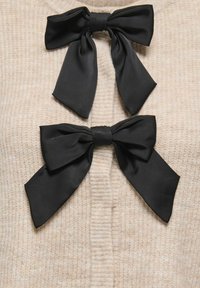 Beżowa dzianina z dwoma dużymi czarnymi satynowymi kokardami przy dekolcie, o gładkiej teksturze i miękkich, prążkowanych detalach.