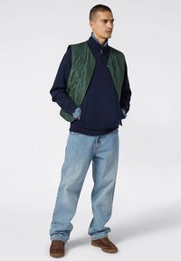 Gilet trapuntato verde sopra una felpa blu navy, abbinato a jeans azzurri e scarpe marroni. Presenta un colletto con zip e una vestibilità comoda.