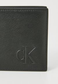 Černá kožená peněženka s texturovaným povrchem, embosovaným logem "cK" a čistým švem podél okrajů. Kompaktní obdélníkový design.
