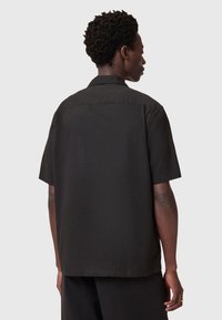 Chemise noire à manches courtes, fabriquée en tissu léger, présente une coupe droite, un empiècement au dos et une texture lisse. Aucun motif ni accent visible.