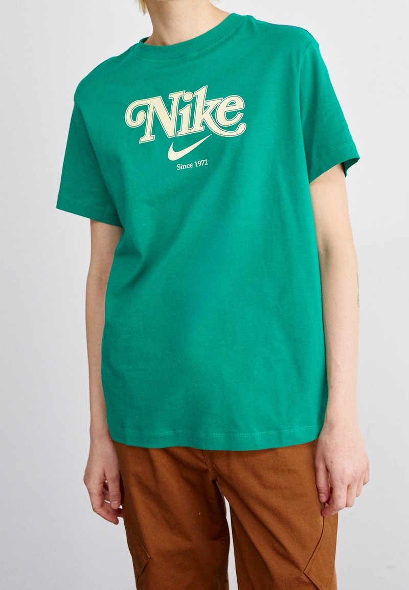 T-shirt en coton vert avec un grand logo vintage "Nike" en crème et le texte "Depuis 1972" en dessous. Manches courtes et col rond.