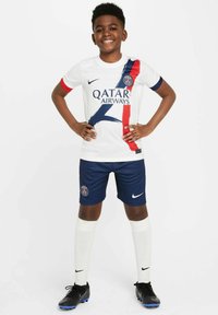 Maillot de football pour les jeunes en blanc avec des accents bleus et rouges, portant le logo "Qatar Airways". Associé à un short et des chaussettes navy, ainsi qu'à des chaussures à crampons noires.