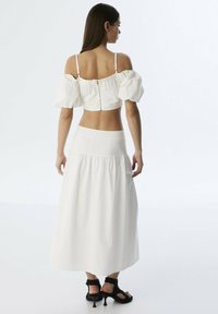 Witte cropped top met pofmouwen; ritssluiting aan de achterkant. In combinatie met een witte, vloeiende maxi rok. Zwarte hakken zichtbaar.