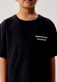 Enfant portant un T-shirt noir avec le texte blanc "AMERICAN VINTAGE" sur la poitrine, sur un fond clair uni.