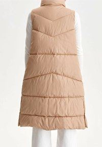 Beige gepolsterte Weste mit hohen Kragen, die ein Zickzackmuster und horizontale Steppabschnitte aufweist, die bis zur Mitte des Oberschenkels reichen.