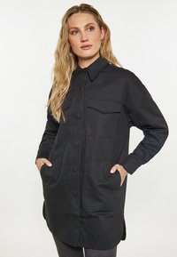 Blondynka ubrana w długi czarny płaszcz zapinany na guziki z czterema dużymi kieszeniami, ręce w bocznych kieszeniach, patrzy lekko w prawo, neutralne tło.