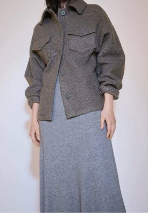 Person trägt eine braune, geknöpfte Wolljacke mit Brusttaschen über einem langen, grauen, strukturierten Kleid.