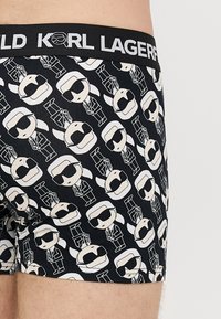 Melni bokseršorti ar atkārtotu baltu zīmējuma rakstu, kurā redzama tēls ar saulesbrillēm un uzvalku, ar melnu jostu, uz kuras uzraksts "KARL LAGERFELD".