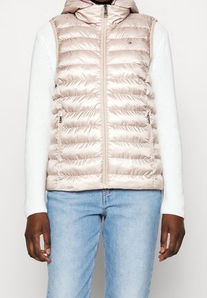 Person trägt eine beige gesteppte Kapuzenweste über einem weißen Strickpullover und hellblauen Jeans, steht mit entspannten Armen an den Seiten.