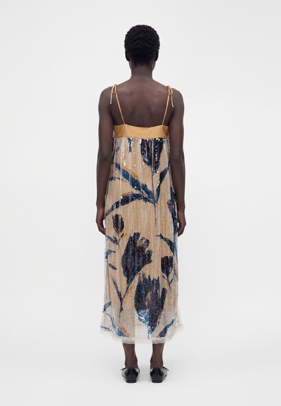 Personne portant une robe midi beige sans manches avec des motifs floraux bleus et des fines bretelles, debout de dos sur fond blanc.