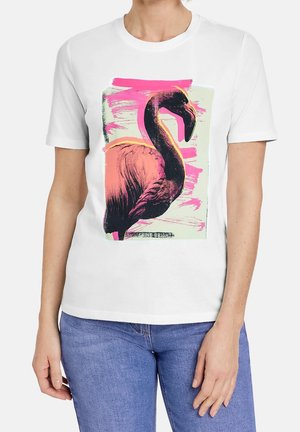 T-shirt blanc à manches courtes avec un motif de flamant rose en tons rose et noir sur des coups de pinceau abstraits roses, porté avec un jean bleu.