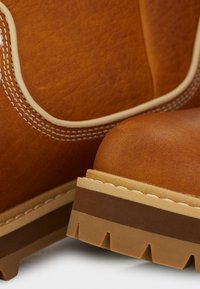 Lähikuva ruskeista nahkasaappaista, joissa on yksityiskohtainen valkoinen ompelu ja paksut kerroskumipohjat valkoista taustaa vasten.