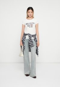 Hvit t-skjorte med "Hollister Co. 1922 So-Cal" trykk, blå rutete skjorte knyttet i livet, lyseblå vide jeans og beige håndveske.