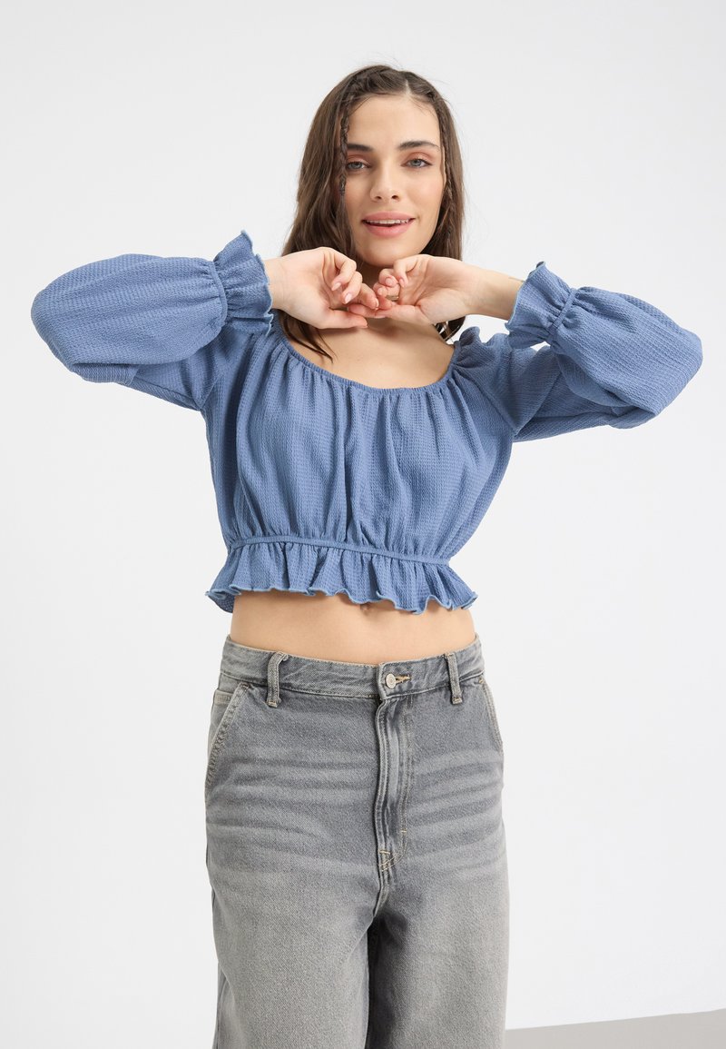 Kék, könnyű textúrájú crop top széles nyakkivágással és puffos ujjal, fodros szegéllyel, magas derekú szürke farmerral párosítva.