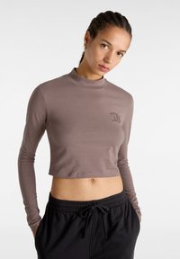 Top corto a maniche lunghe in tessuto marrone chiaro con collo alto e logo VANS ricamato sul petto; abbinato a pantaloni neri con cordino.