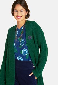 Lächelnde Frau in grünem strukturiertem Cardigan, blauer Bluse mit Blumenmuster, geschichteten Perlenhalsketten, goldenen Ohrringen mit Tropfen und dunkelblauen Hosen mit Knöpfen.