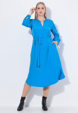 Abito blu al ginocchio con scollo a V, maniche lunghe e cintura a fiocco. Presenta una silhouette fluida e tasche laterali. Indossato con stivali neri alla caviglia.