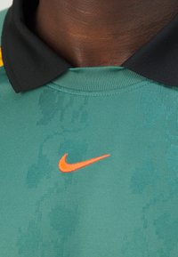 Polo-shirt de sport vert avec un motif texturé, col noir et logo Nike swoosh orange sur le côté gauche.