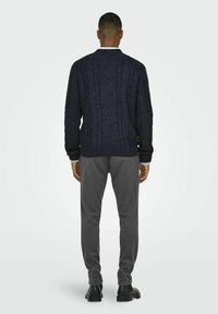 Pull en laine navy à mailles torsadées avec un col haut, associé à un pantalon gris et des chaussures noires, présenté de dos.