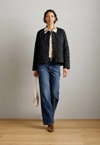 Giacca nera trapuntata con colletto rotondo, abbinata a una maglietta a fantasia e jeans larghi blu. Gli accessori includono una borsa chiara e stivali marroni.