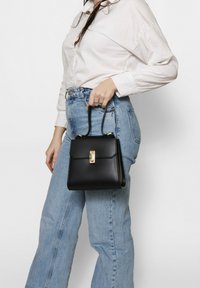 Borsa a mano in pelle nera con forma strutturata, dotata di una chiusura dorata e di un manico corto, tenuta da una persona che indossa jeans azzurri e una camicia bianca.