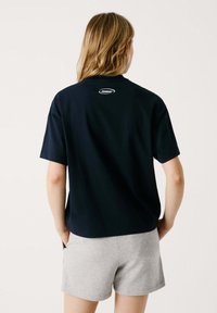 Meripaita lyhyillä hihoilla, rentoa istuvuutta, tummansininen, jossa on logopatchi takana ja pehmeää puuvillamateriaalia. Yhdistetty harmaisiin shortseihin.
