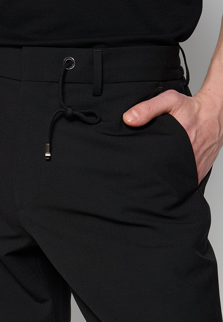 Gros plan sur une main dans la poche droite d'un pantalon noir avec une taille à cordon de serrage dotée de cordons à embouts en métal.