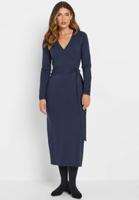 Marineblaues Wickelkleid mit langen Ärmeln, V-Ausschnitt und Bindegürtel. Glatter Stoff; knöchellang. Kombiniert mit schwarzen Strumpfhosen und Schuhen.
