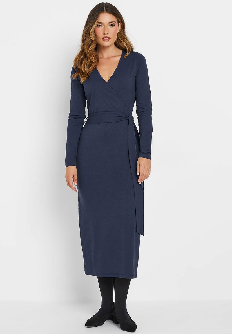 Marineblaues Wickelkleid mit langen Ärmeln, V-Ausschnitt und Bindegürtel. Glatter Stoff; knöchellang. Kombiniert mit schwarzen Strumpfhosen und Schuhen.