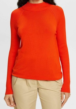 Femme portant un pull à manches longues côtelé de couleur orange vif avec un pantalon beige sur un fond clair uni.
