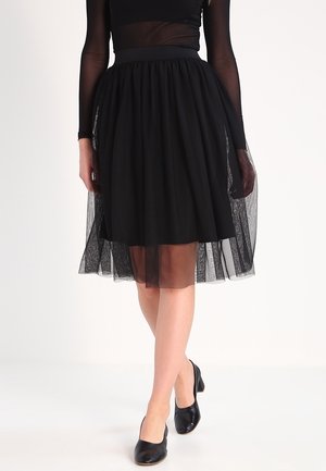 Mujer con falda negra de tul hasta la rodilla y blusa negra transparente de manga larga, con zapatos negros de tacón bajo, de pie con las piernas cruzadas.