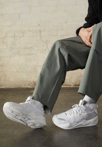 Vitaidiga idrottsskor i vitt med en ovandel av mesh, texturerade överlager och gummisulor. Bärs med ljusgrå strumpor och löst sittande gröna byxor.