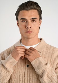 Beige kabelstickad tröja med krage, knapplist och ribbad manschett. Modellen justerar kragen med båda händerna och bär en vit skjorta under.