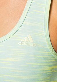 Jasnozielony sportowy top na ramiączkach z gradientowym wzorem, wykonany z gładkiego materiału, z białym logo Adidas na lewej części klatki piersiowej.