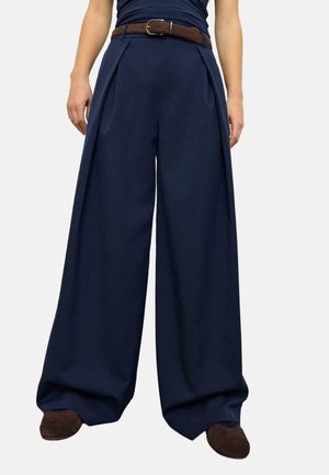 Persona che indossa pantaloni largi blu navy con pieghe, cintura marrone, scarpe marrone scuro e una maglia blu navy infilata, in piedi con le mani rilassate.