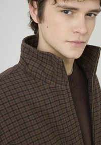 Veste zippée marron à motifs avec col montant, présentant un motif géométrique dans des tons plus foncés, portée sur un pull marron.