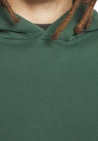 Närbild av en person som bär en grön huva med bruna dreadlocks som vilar på axlarna, visar nacke och övre bröstområde.