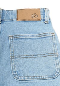 Vaaleansiniset farkut, joissa on ruskea nahkapatchi, jossa on kuvia. Taskuissa ja vyötäröllä on näkyvissä ompelutiedot.